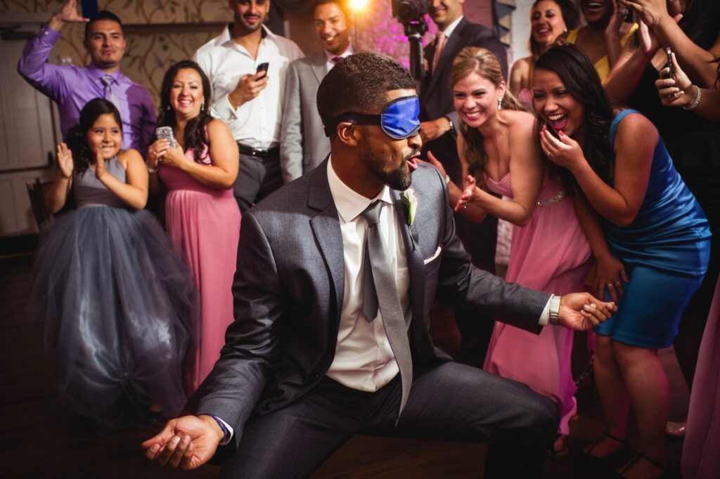 Man in a blindfold dancing at a Bronx Zoo Stone Mill wedding reception while guests cheer around him.