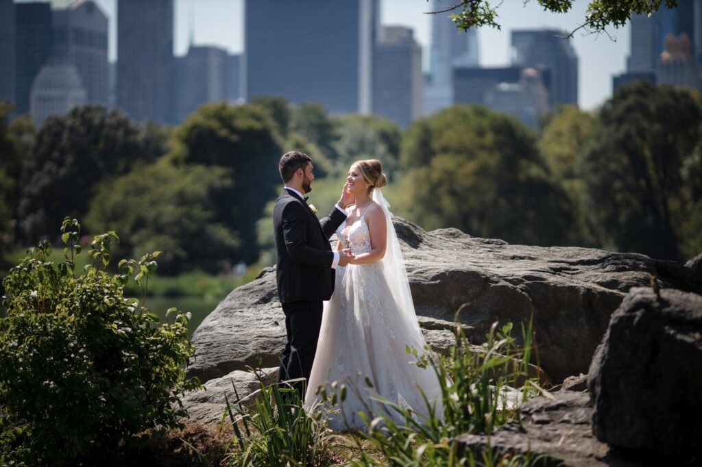 Bride and groom holding hands on a rock in Central Park with the city skyline in the background.