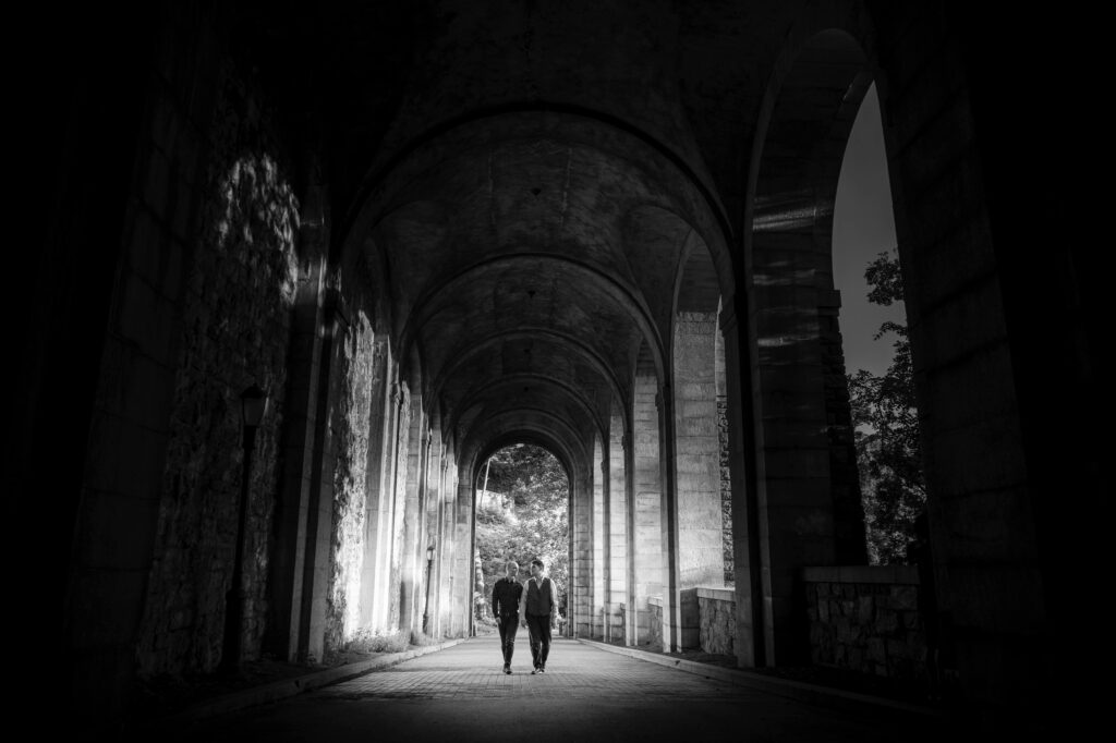 Couple walking through a shaded archway into the light for their Fort Tryon Park engagement photos, featuring two grooms.