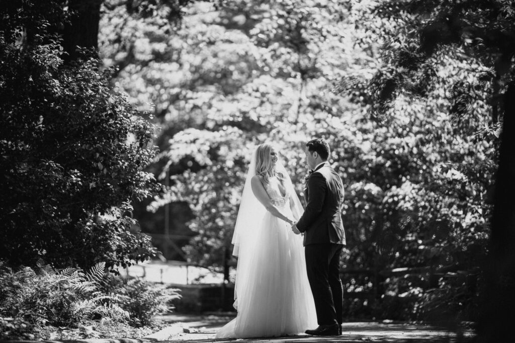 A bride and groom holding hands outdoors at the Bronx Zoo Stone Mill, surrounded by trees bathed in sunlight.