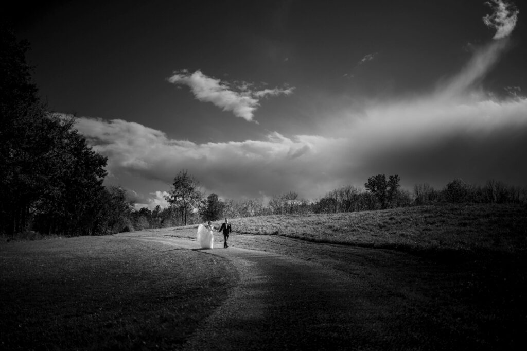 A couple holding hands and walking down a winding road in a serene landscape, with dramatic clouds above, evoking the enchantment of a Meadow Ridge on the Hudson wedding.