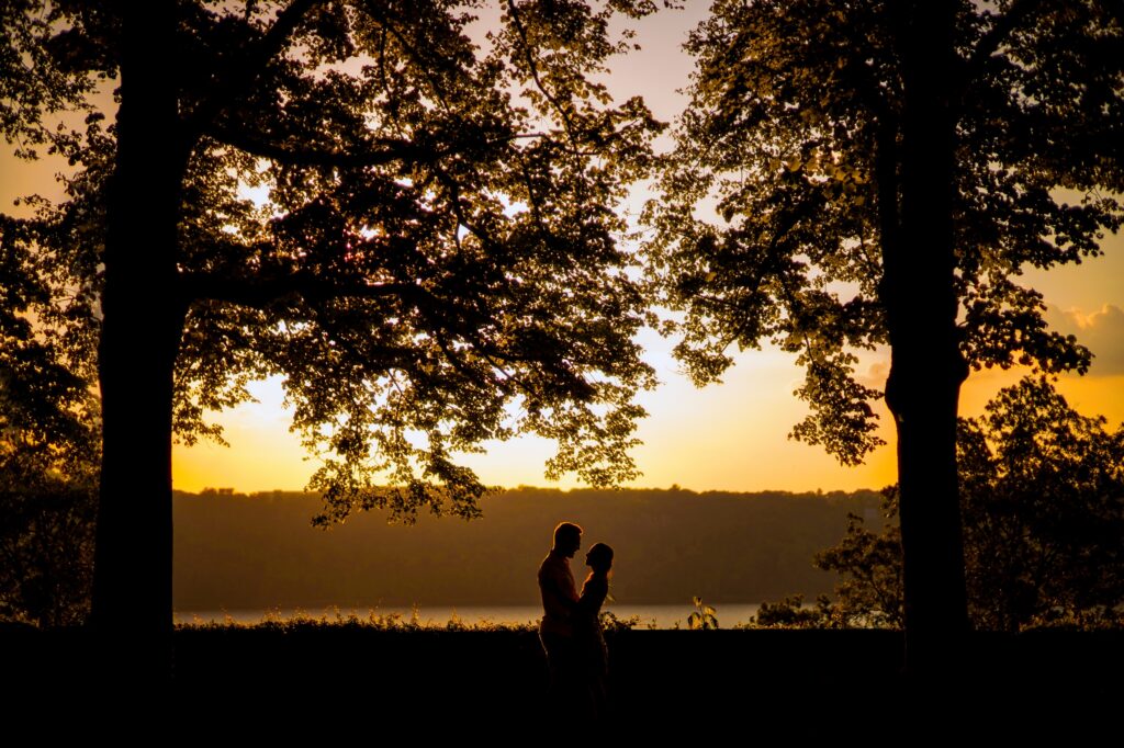 Two silhouetted individuals standing between trees in Fort Tryon Park, with a sunset over a lake in the background.