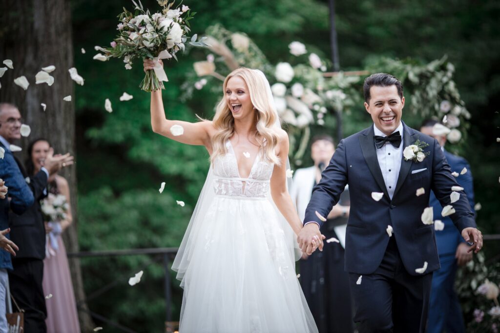 A joyful bride and groom walk hand in hand at the Bronx Zoo Stone Mill as wedding guests throw petals to celebrate their union.