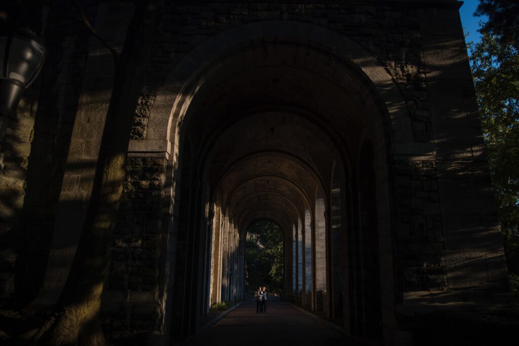Two grooms walk through a shaded corridor with arched ceilings and columns in Fort Tryon Park, illuminated by sunlight at the far end.