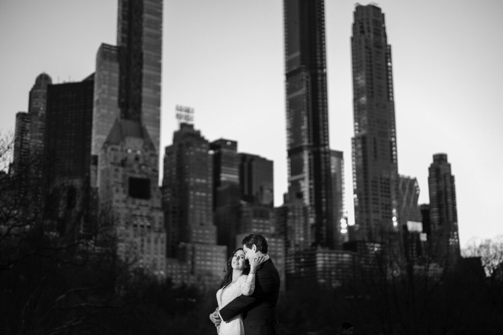 A couple embracing in Central Park with city skyscrapers in the background.