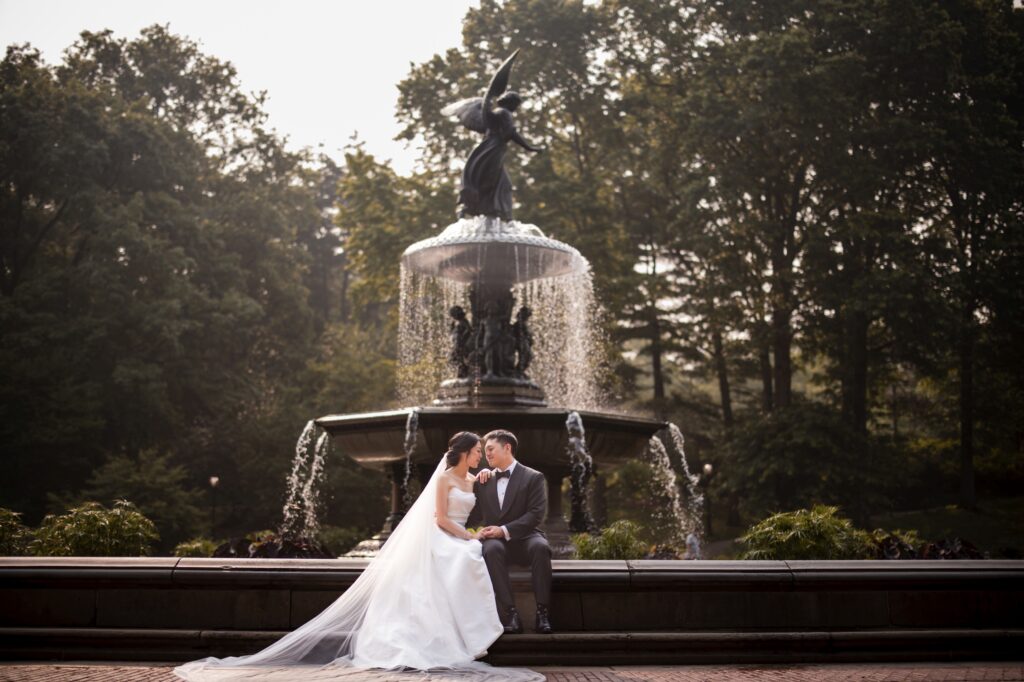 A bride and groom share a kiss by a fountain with a statue in Central Park.