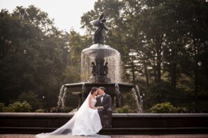 A bride and groom share a kiss by a fountain with a statue in Central Park.