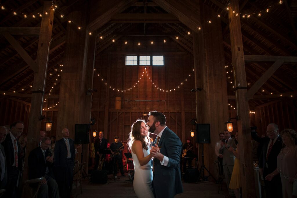 A couple shares their first dance at a Stonover Farm Wedding in a rustic barn illuminated by string lights as guests look on.