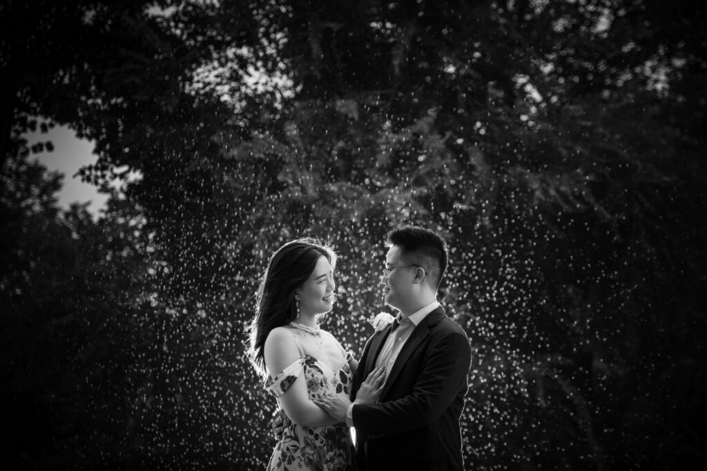 A monochrome image capturing a tender moment between a couple, with the man in a suit and the woman in a floral dress, surrounded by a magical spray of water droplets backlit by sunlight, creating a dreamlike atmosphere.