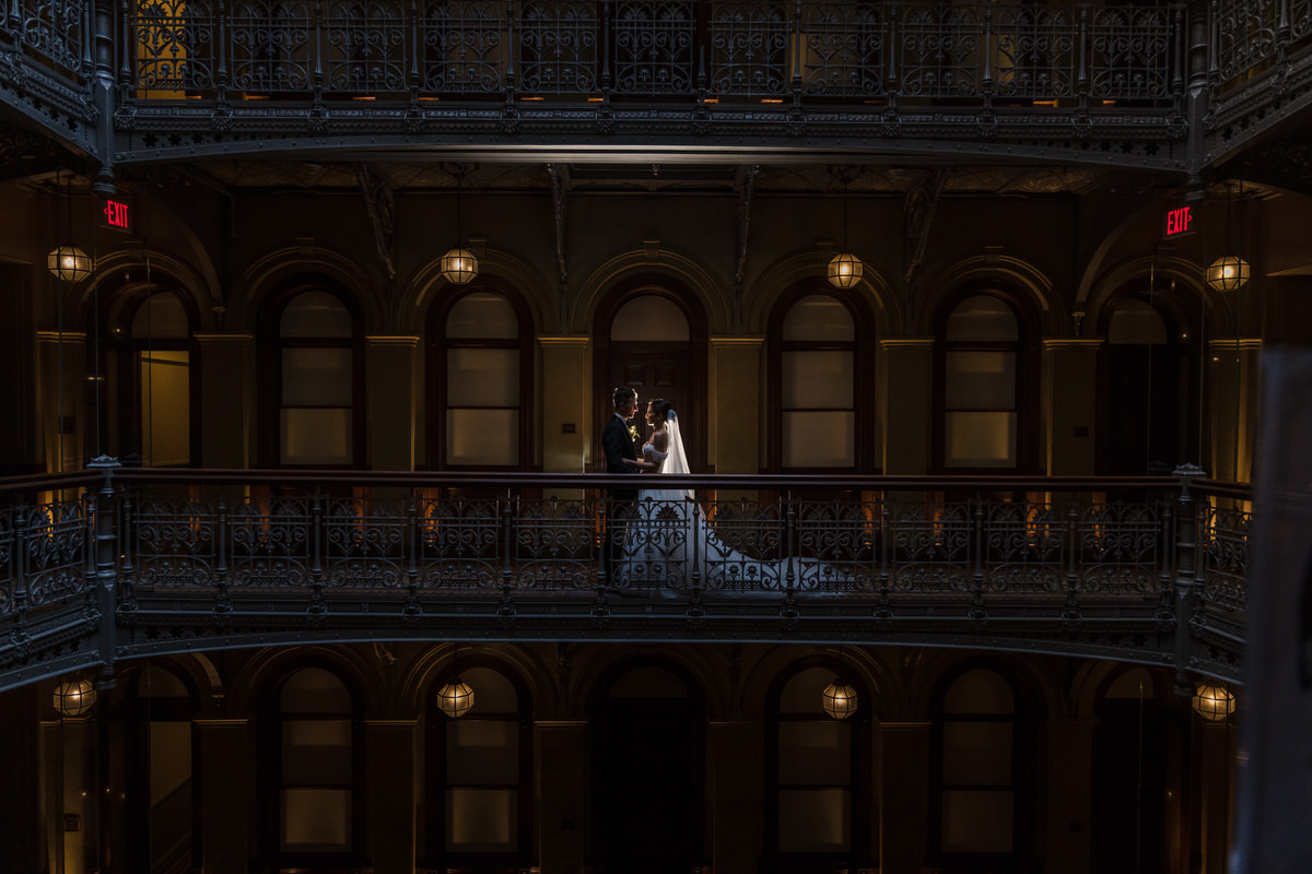 Dramatic lighting highlights a bride and groom standing intimately on a balcony, with the grandeur of a historic, opulently detailed building surrounding them, emphasizing the magnificence of the venue and the intimacy of their union.