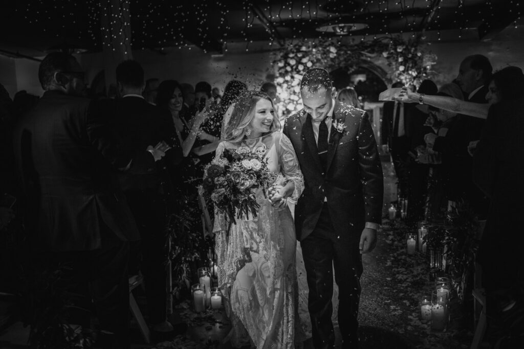 Black and white image of a smiling bride and groom walking hand in hand, surrounded by their guests throwing confetti on their wedding day