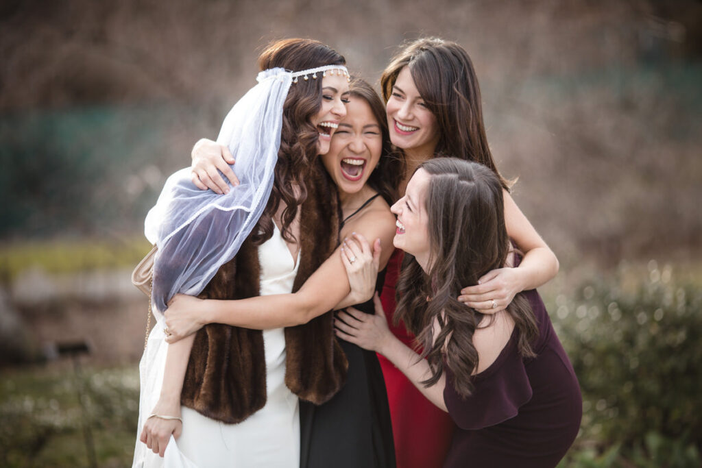 Bride laughing joyously while being embraced by bridesmaids in an outdoor setting