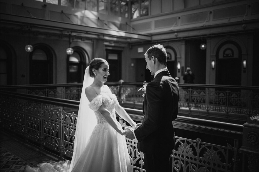Black and white photo of a joyful bride in an intricate lace wedding gown, sharing a laugh with the groom in a classic tuxedo, inside an elegant venue with ornate railings and warm lighting from overhead lamps.