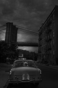 A romantic black and white shot of a newlywed couple sharing a private moment in the back of a vintage taxi, with the iconic Brooklyn Bridge in the background, under a dramatic sky at dusk.