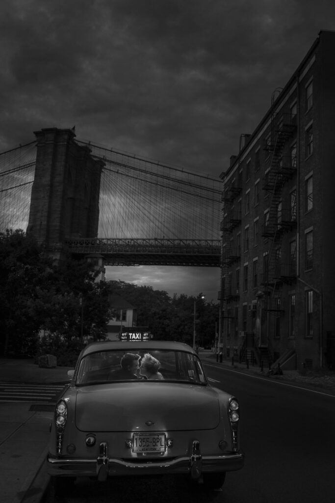 A romantic black and white shot of a newlywed couple sharing a private moment in the back of a vintage taxi, with the iconic Brooklyn Bridge in the background, under a dramatic sky at dusk.