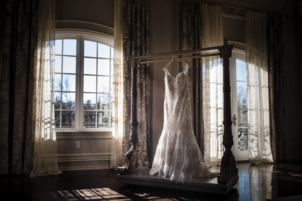 A Park Chateau wedding dress hanging on a display in a sunlit room with elegant drapes.