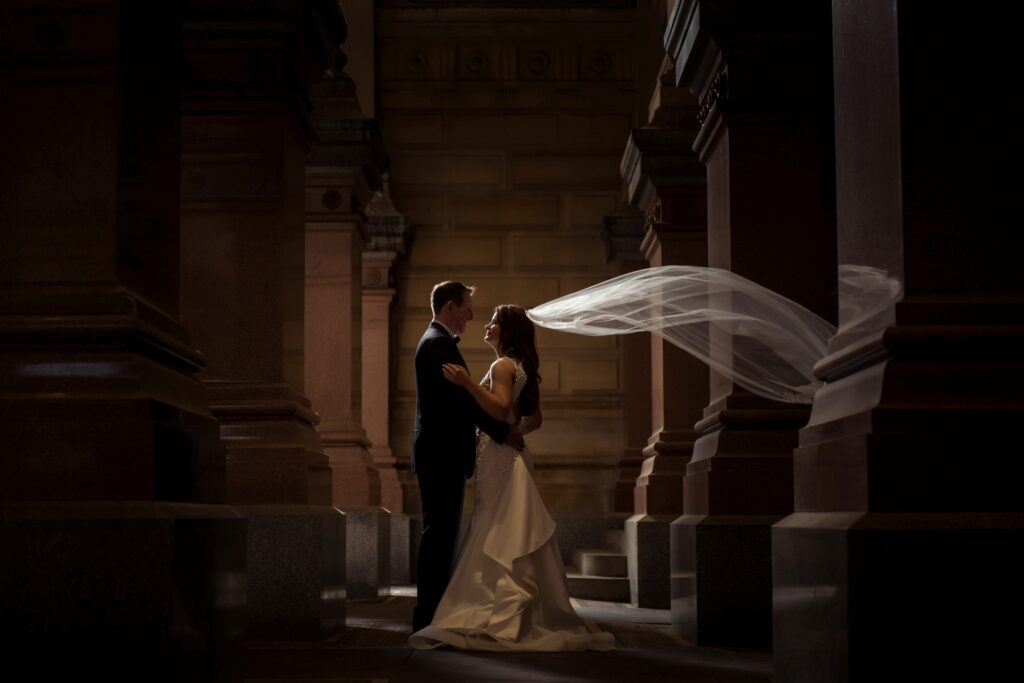 A bride and groom sharing a moment in a dimly lit, architectural setting beneath Philadelphia PA City Hall, with the bride's veil caught in a gentle breeze.