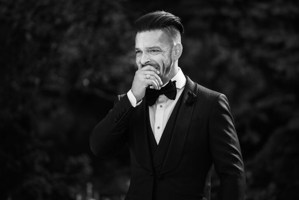 A monochrome image of a man in a tuxedo smiling and touching his chin, standing outdoors at Crystal Plaza with trees in the background.
