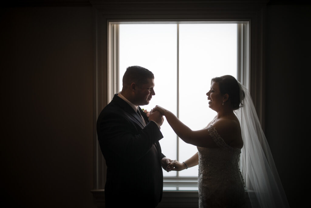 A backlit bride and groom holding hands by a window at their Park Chateau wedding, creating a silhouette effect.