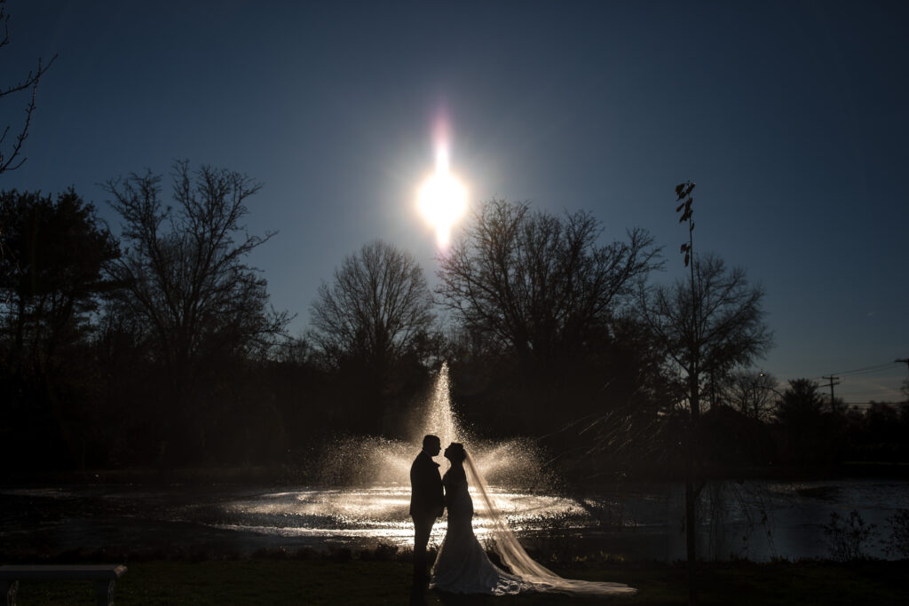 A couple in wedding attire standing by a pond with a fountain at Park Chateau, backlit by the sun creating silhouettes.