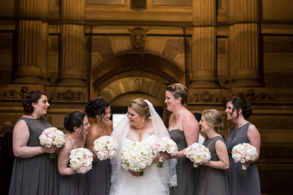 A bride and her bridesmaids smiling and holding bouquets, captured in an elegant, architectural setting at Philadelphia PA City Hall.