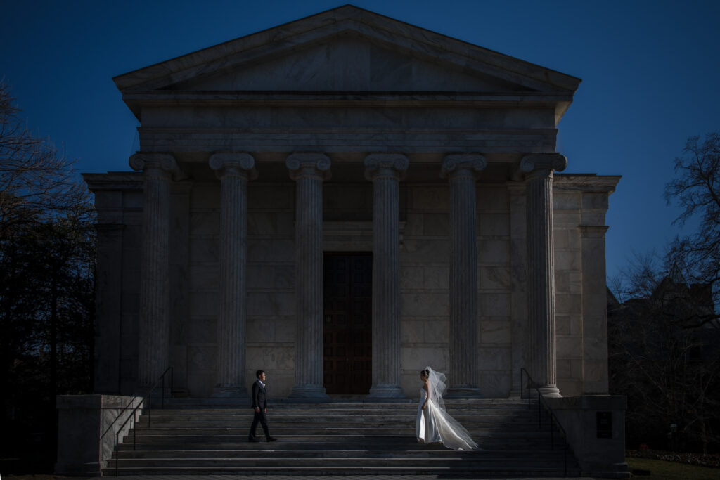 A bride and groom standing apart on the steps of a neoclassical building with tall columns under a clear sky at Princeton University.