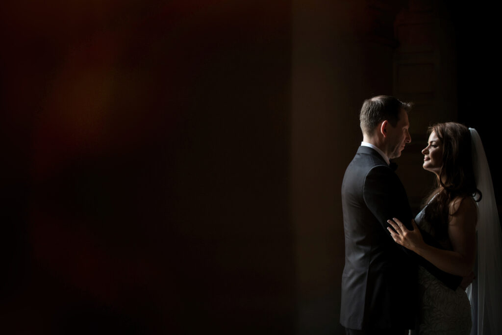 A bride and groom share an intimate moment underneath Philadelphia's City Hall in a dimly lit room with a soft spotlight.