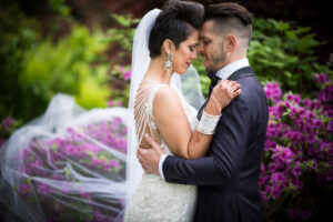 A bride and groom embracing tenderly at a Crystal Plaza wedding among blooming pink flowers, with the bride's veil trailing and detailed dress visible.