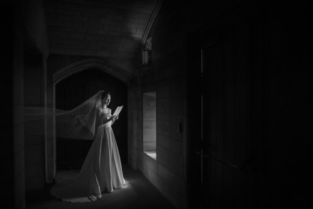 A bride in a white gown reading a letter by the light of a window in a dimly lit corridor at Princeton University.