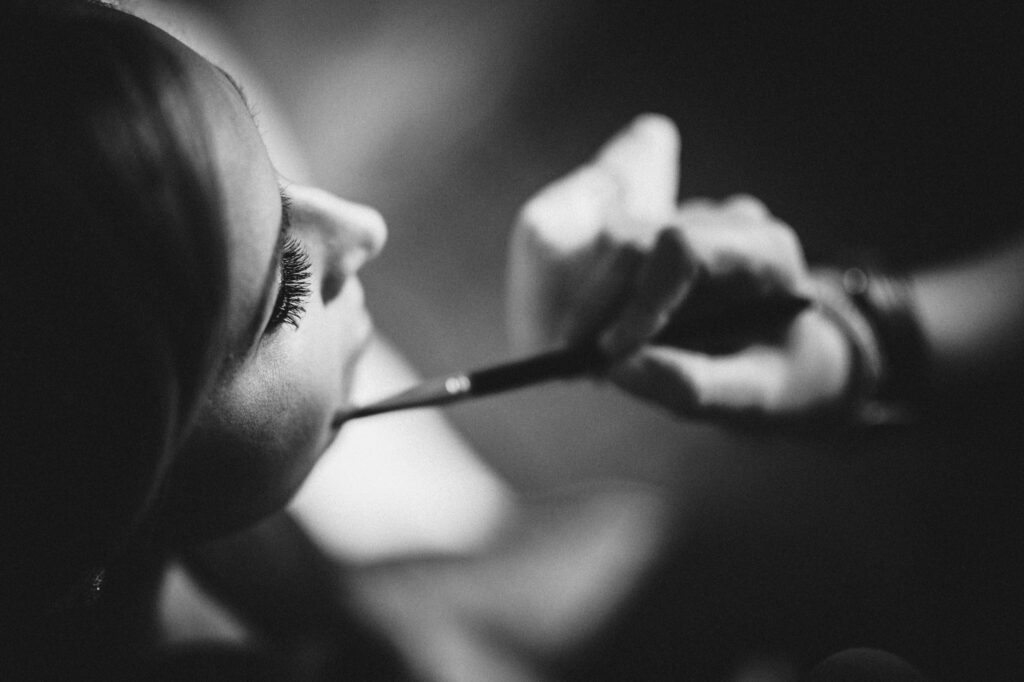 Black and white photo of a woman having makeup applied to her lips by another person using a brush at a Maritime Parc wedding.