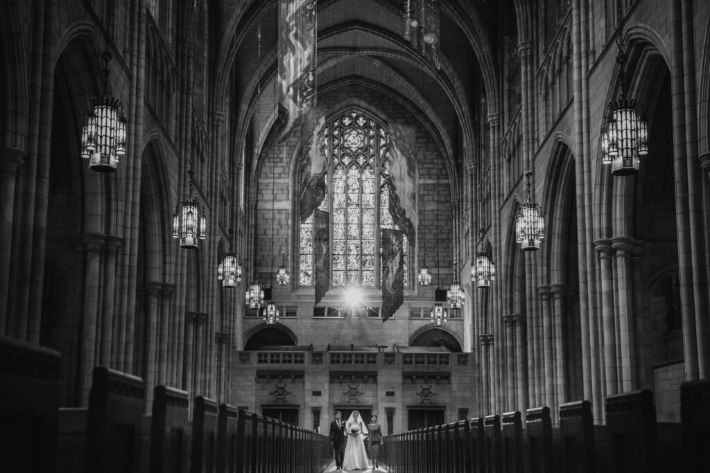 Bride, accompanied by her mom and dad, walks down the aisle of Princeton University's chapel, illuminated by the light from a large stained glass window, with rows of pews on either side.