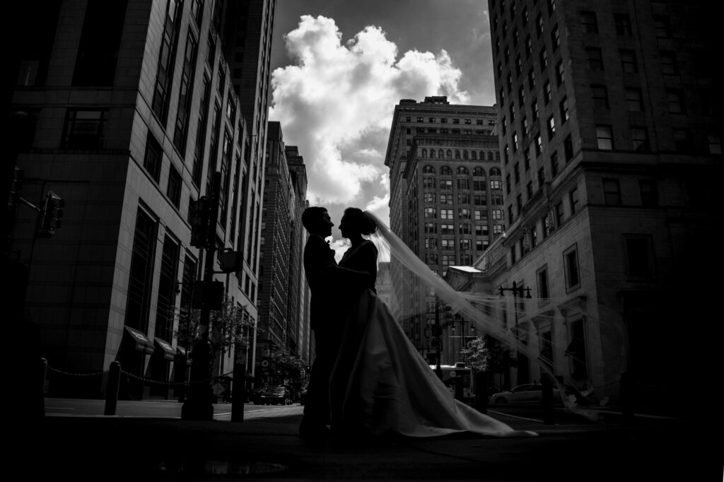 A silhouetted couple sharing a romantic kiss on a city street near Philadelphia's City Hall, framed against a backdrop of towering buildings and a dramatic sky.