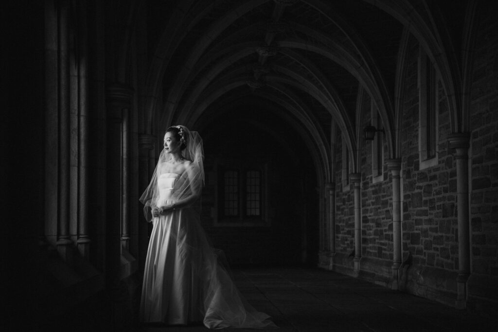 A bride standing alone in a gothic corridor at Princeton University, lit by soft light from the side.