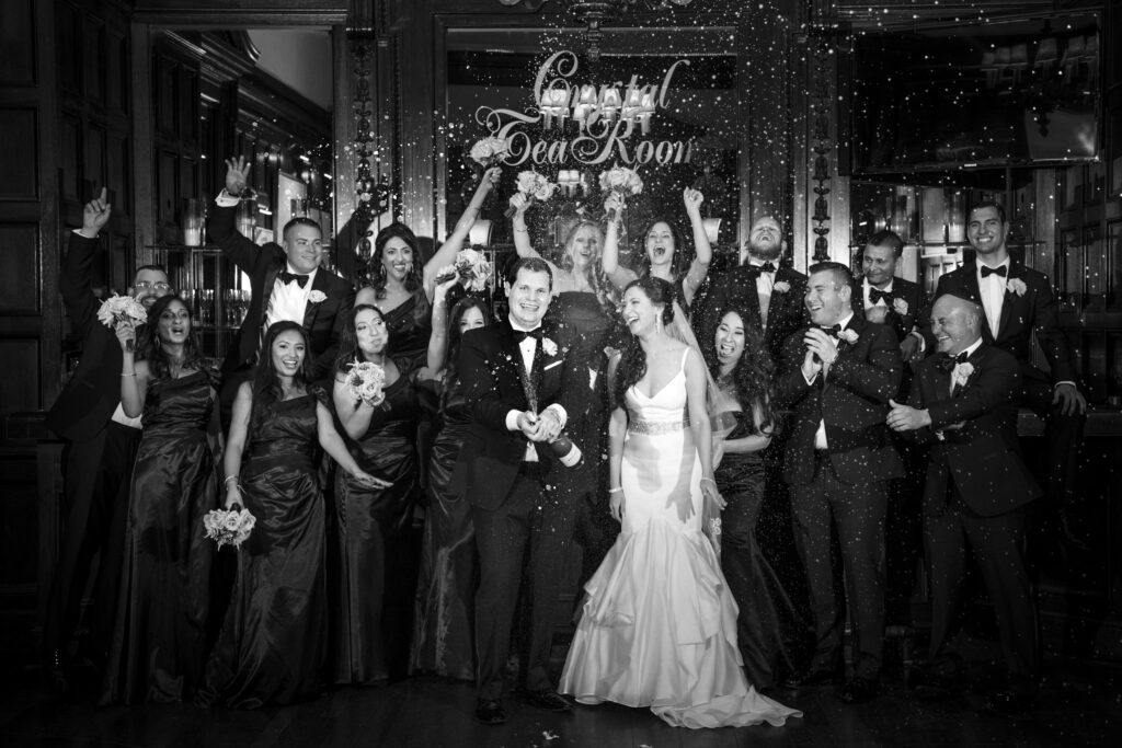 A black and white photo of a joyful wedding party celebrating with the bride and groom in a room with "Crystal Tea Room Philly" sign, throwing confetti.