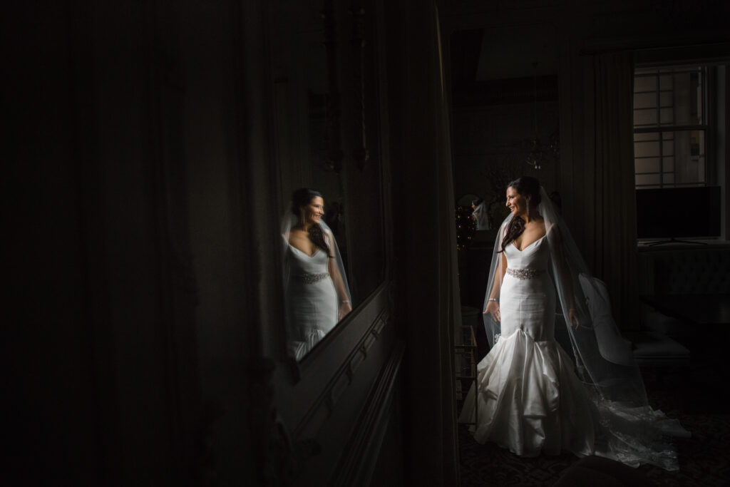 A bride in a white gown stands by a mirror in the dimly lit Crystal Tea Room, her reflection visible as she looks to the side.