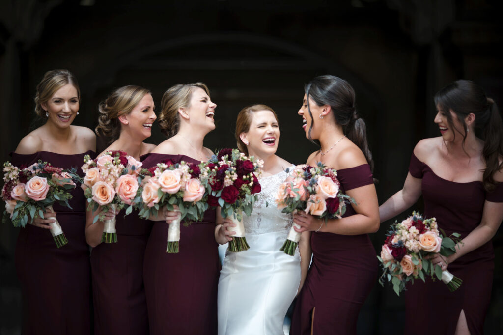 A bride laughing joyfully surrounded by her bridesmaids at Philadelphia City Hall, who are dressed in burgundy gowns and holding bouquets of flowers.