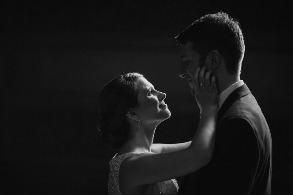 A black and white photo of a woman tenderly holding a man's face as they gaze into each other's eyes in front of Philadelphia's City Hall after their wedding.