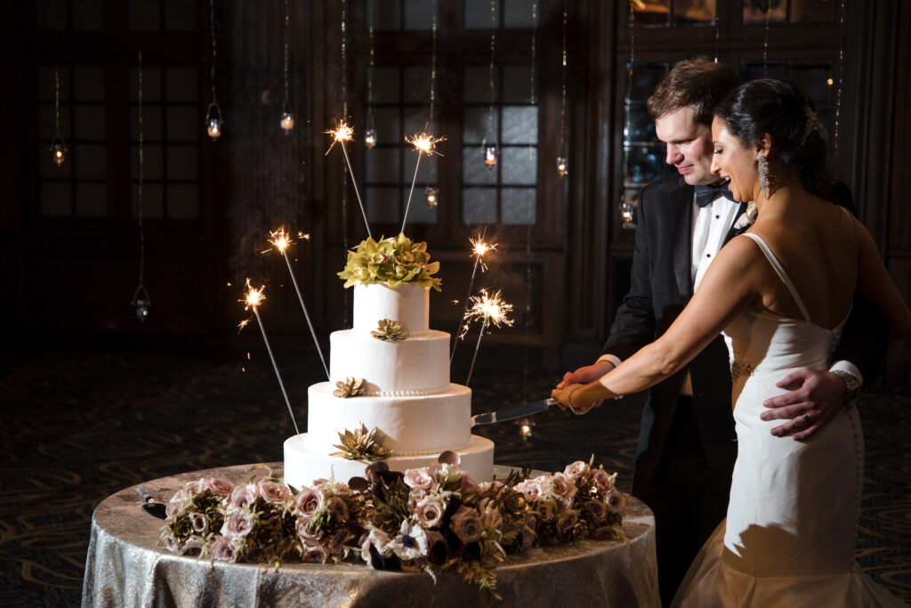 A bride and groom cutting their wedding cake, adorned with sparklers and surrounded by floral arrangements, in the Crystal Tea Room during a dimly lit Philly wedding.