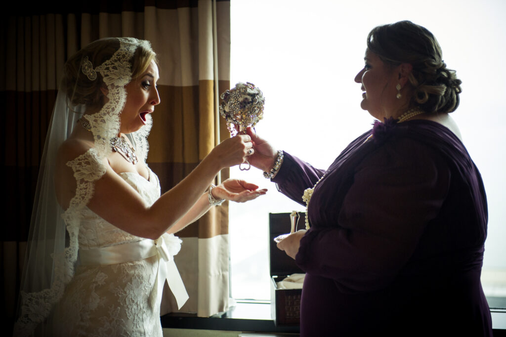 A bride in a lace dress excitedly shows a bejeweled bouquet to an older woman in a purple dress by a window at a Maritime Parc wedding.