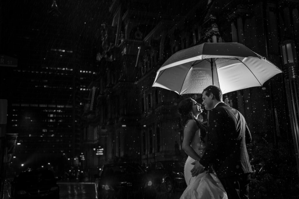 A couple shares a romantic kiss under a white umbrella on a rainy night outside the Crystal Tea Room in Philly, with streetlights and raindrops visible.