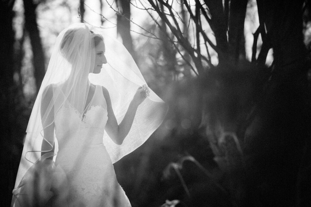 A bride in a white dress and veil looks to the side, standing in a wooded area with soft, natural light filtering through the trees during her Crystal Plaza wedding.