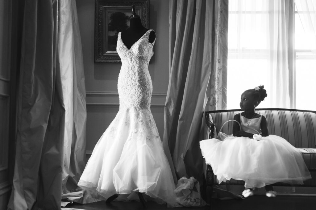 A young girl in a white dress sits on a sofa at the Park Savoy, looking at a wedding dress displayed on a mannequin by a window, in a monochrome setting.
