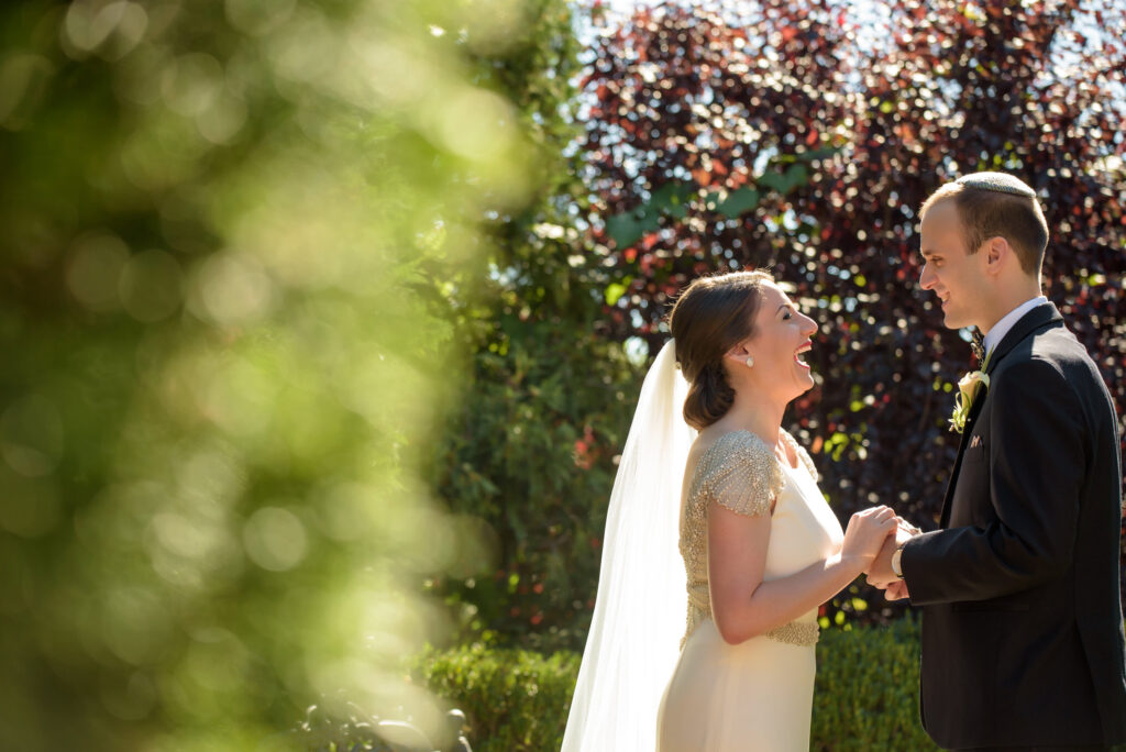 A bride and groom holding hands and laughing on a sunny day at a Park Savoy wedding, framed by green foliage and a darker foliage backdrop.