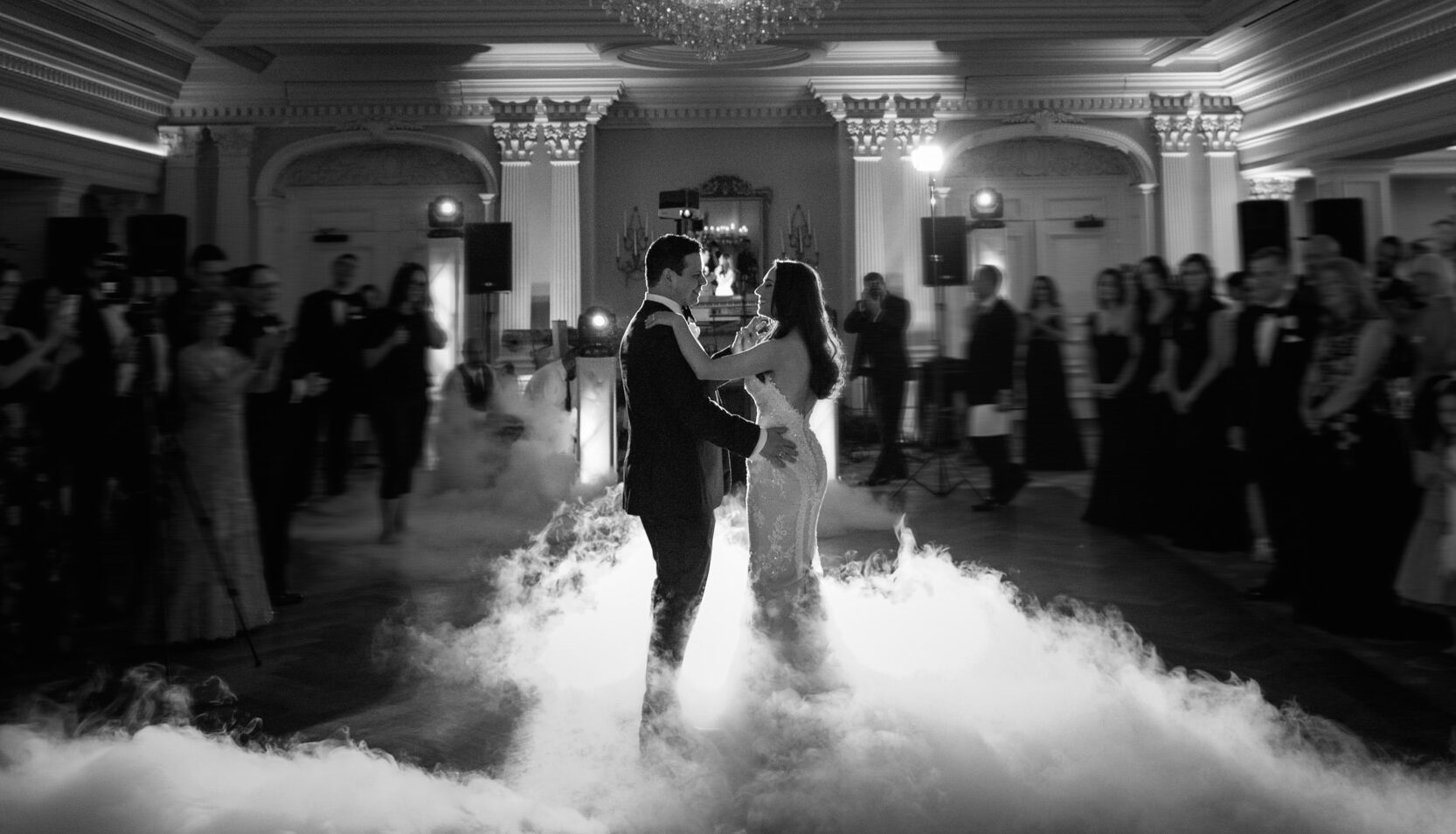 A couple shares their first dance on a smoke-filled dance floor under a grand chandelier at their Park Savoy wedding, surrounded by guests in an elegant ballroom.