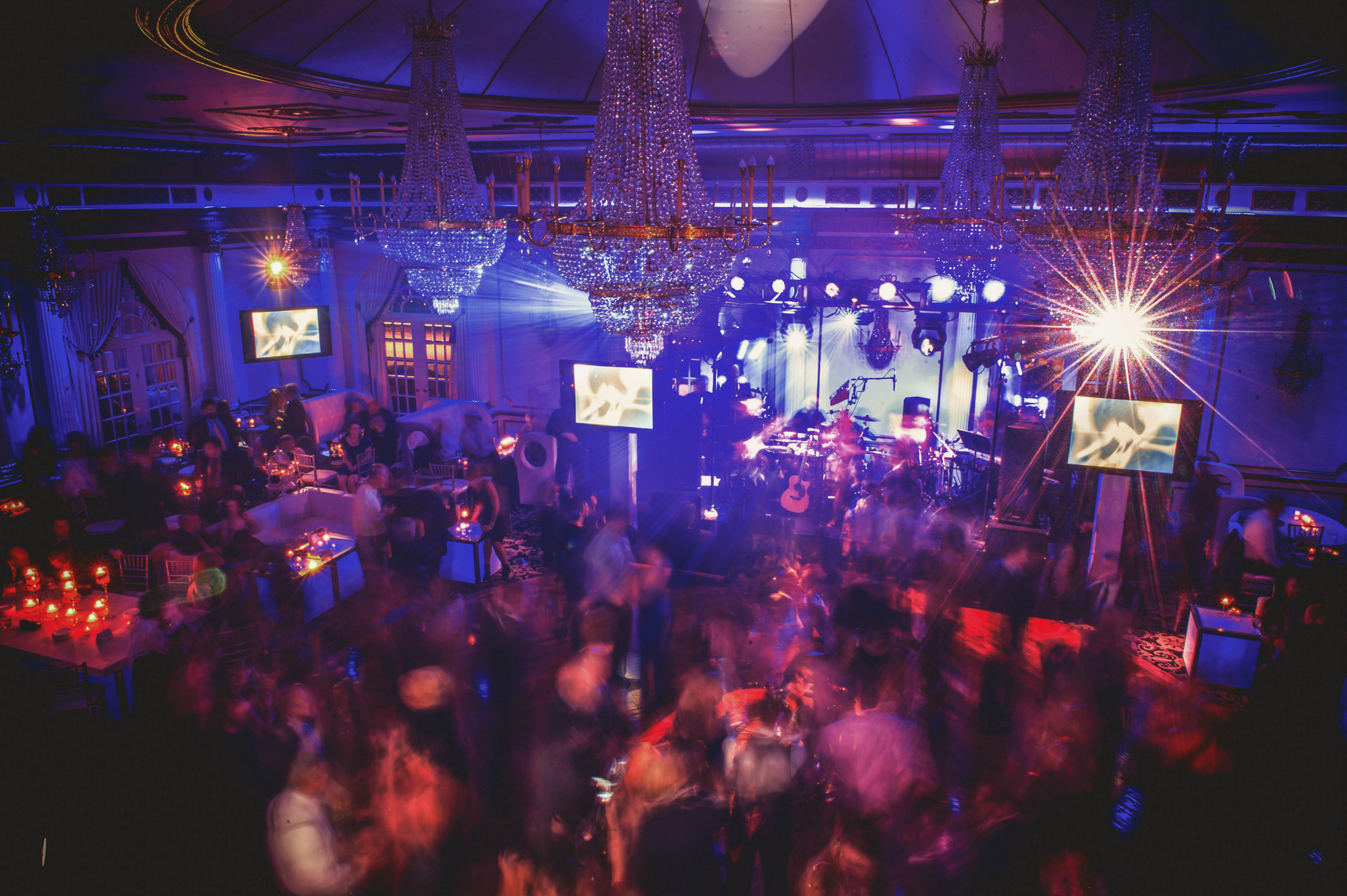 Crystal Plaza wedding venue with bluish haze and chandeliers overhead; crowd dancing with motion blur, band performing on stage lit by vibrant lights.