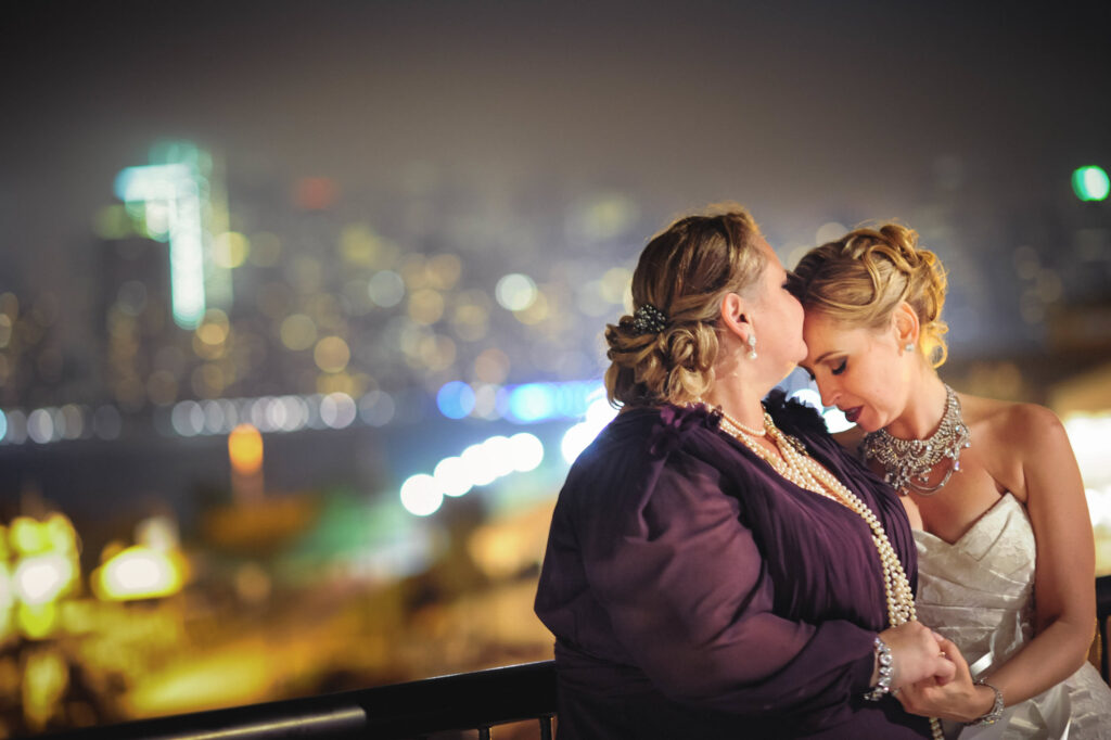 Two women embracing and kissing on a balcony at their Maritime Parc wedding with a blurred city skyline at night in the background.