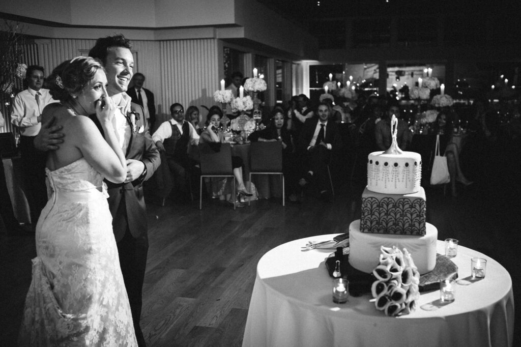 A bride and groom laugh together beside their wedding cake in a Maritime Parc reception hall filled with guests.