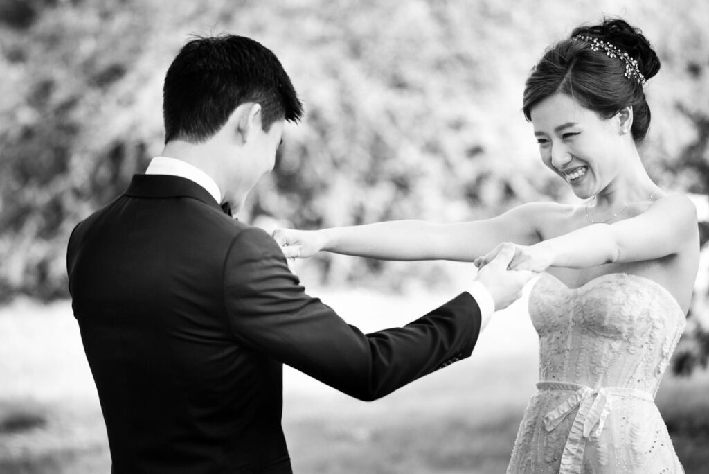 A monochrome image of a smiling bride playfully dancing with her groom outdoors, with trees softly blurred in the background.