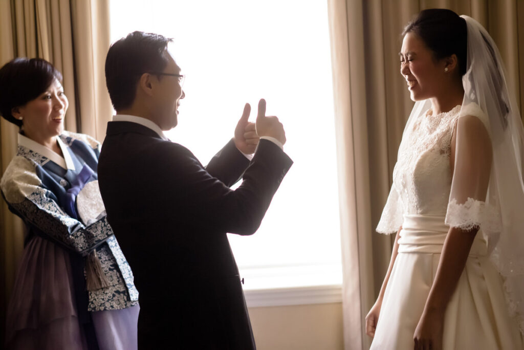 A father, playfully gesturing a thumbs-up, receives an adoring glance from his bride in a luminous room while a woman in traditional Korean attire smiles on.