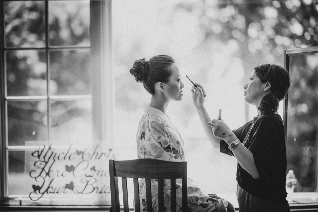 A black and white photo of a bride sitting by a window, having makeup applied, with a sign reading “Here Comes Your Bride” in the foreground.